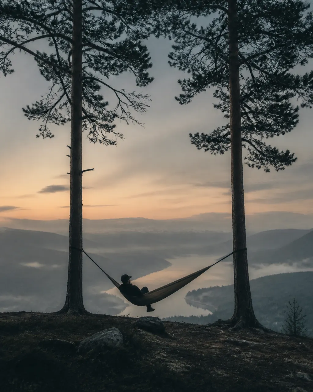 A man resting in a hammock between pines overlooking a misty valley at dusk