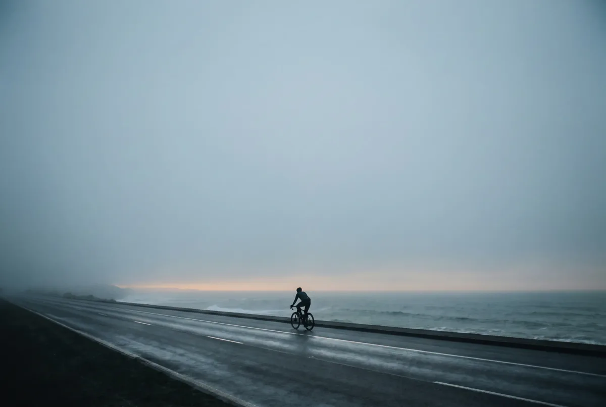 A lone cyclist riding a coastal road into low evening fog